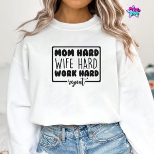 May include: White sweatshirt with the text "MOM HARD, WIFE HARD, WORK HARD, repeat" in a black rectangular box. The sweatshirt is a crew neck style and is paired with blue denim jeans.
