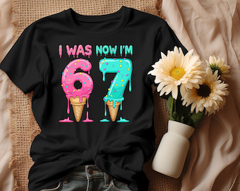 May include: Black t-shirt with the text "I WAS NOW I'M" above the numbers "67". The numbers are designed as ice cream cones, with pink and blue dripping ice cream and sprinkles. A bouquet of yellow flowers in a white vase is to the right.