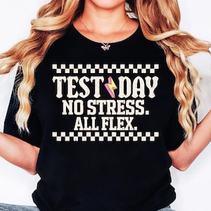 May include: Black t-shirt with a white checkered border and the text "TEST DAY NO STRESS. ALL FLEX." in white with a pink and yellow lightning bolt graphic.