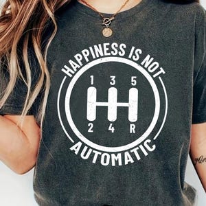 May include: Dark grey t-shirt featuring a white gear shift graphic with the text "HAPPINESS IS NOT AUTOMATIC". The gear shift design includes the numbers 1, 2, 3, 4, 5, and R.