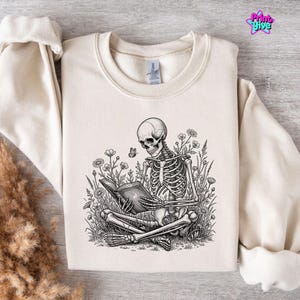 Puede incluir: Sudadera color crema con una ilustración detallada en blanco y negro de un esqueleto leyendo un libro en un campo de flores. También hay una mariposa en el diseño. La marca "Print & Give" es visible.