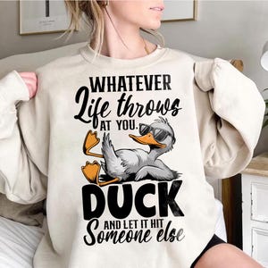 May include: A cream colored sweatshirt with the text "Whatever life throws at you, duck and let it hit someone else" and a cartoon duck wearing sunglasses.