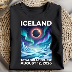 May include: Black t-shirt with a colourful graphic depicting a total solar eclipse over an icy landscape with aurora borealis. The word "ICELAND" is at the top, with "TOTAL SOLAR ECLIPSE AUGUST 12, 2026" below the graphic.