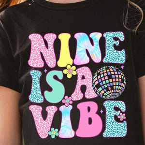 以下が含まれることがあります： 「NINE IS A VIBE」というフレーズがカラフルなレトロスタイルでプリントされた黒いTシャツ。デザインには、ディスコボール、花、タイダイやヒョウ柄などの様々なパターンが含まれています。