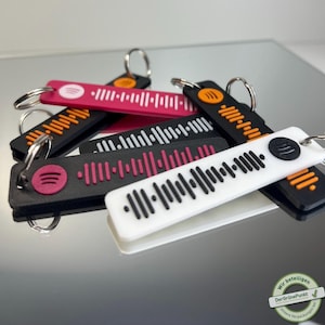 Könnte beinhalten: Ein Set mit sechs Schlüsselanhängern mit verschiedenen farbigen Spotify-Barcodes. Die Schlüsselanhänger sind aus schwarzem, weißem, orangefarbenem und rosafarbenem Kunststoff. Jeder Schlüsselanhänger hat einen kleinen Metallring zum Anbringen an Schlüssel.