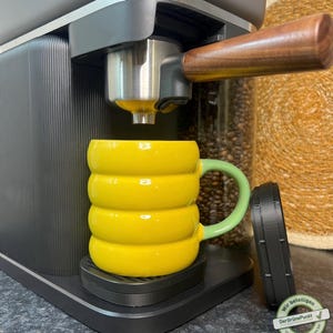 Könnte beinhalten: Eine schwarze Espressomaschine mit einem Holzgriff und einer gelben Tasse mit einem grünen Griff. Die Tasse steht auf einer schwarzen Plattform.
