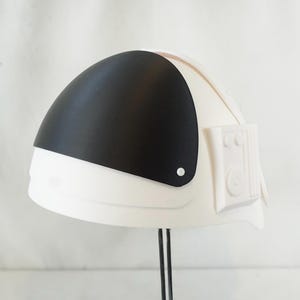 May include: A white astronaut helmet with a black visor. The helmet has a rectangular detail on the side with circular and rectangular features. The helmet is on a black stand.