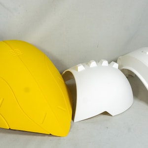 Puede incluir: Conjunto de tres componentes de pl&aacute;stico sin pintar, probablemente para un veh&iacute;culo o disfraz. El conjunto incluye una pieza grande amarilla brillante en forma de casco y dos piezas blancas.