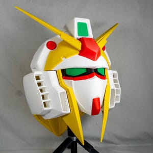 Puede incluir: Una cabeza de robot blanca, amarilla, roja y verde con &aacute;ngulos agudos y detalles. La cabeza del robot tiene un dise&ntilde;o distintivo con una cara prominente, rasgos angulares y un par de antenas amarillas. La cabeza del robot est&aacute; montada en un tr&iacute;pode negro.