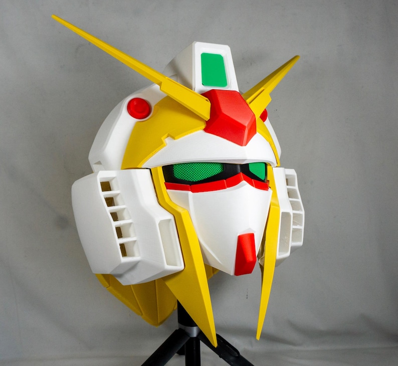 Puede incluir: Una cabeza de robot blanca, amarilla, roja y verde con &aacute;ngulos agudos y detalles. La cabeza del robot tiene un dise&ntilde;o distintivo con una cara prominente, rasgos angulares y un par de antenas amarillas. La cabeza del robot est&aacute; montada en un tr&iacute;pode negro.