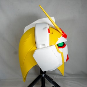 Puede incluir: Vista lateral de un casco de robot amarillo, blanco, rojo y verde. El casco tiene una cresta amarilla y una visera verde. El casco est&aacute; montado en un tr&iacute;pode negro.