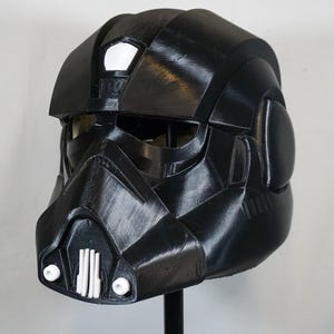 Può includere: Un casco nero dal design distintivo, con una visiera, un elemento prominente per il mento e vari dettagli angolari. Il casco ha una finitura lucida e sembra essere una replica o un elemento di costume. Il casco è esposto su un supporto.