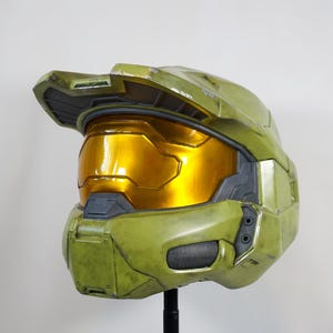 May include: A green and gold helmet, possibly a prop or collectible. The helmet has a gold visor and a detailed design, likely from a science fiction or video game. The item is displayed against a plain background.