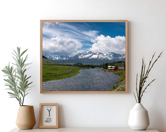 Op de afbeelding: Een ingelijste foto van een rivier die door een vallei slingert met besneeuwde bergen op de achtergrond. De lucht is blauw met witte wolken. Er zijn een paar huizen in de verte.