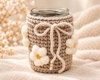 May include: A clear glass jar encased in a beige crocheted cozy featuring a decorative bow tie and three-dimensional white flower appliqués with yellow centers. The cozy is finished with two small wooden beads at the ends of the drawstring ties.