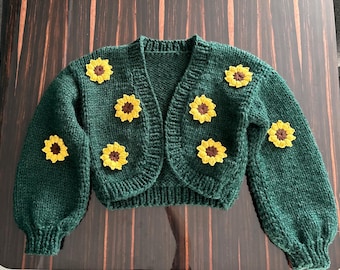 May include: A dark green, cropped cardigan sweater adorned with yellow and brown sunflower appliqués. The knitted sweater features a V-neck and long sleeves, with the sunflower details evenly spaced across the front and sleeves.