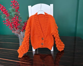 May include: Orange knitted cardigan with a V-neck and button closure. The cardigan has puffed sleeves with a textured knit.