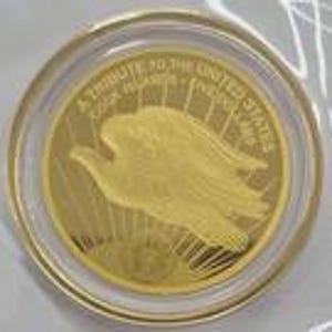 Op de afbeelding: Een goudkleurige munt met een gedetailleerd adelaarsontwerp in een transparante beschermhoes. De munt heeft de inscriptie "A TRIBUTE TO THE UNITED STATES" en andere tekst. De munt is rond en lijkt van metaal te zijn.