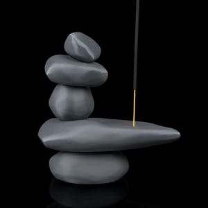 May include: A gray stone incense holder with a stack of smooth, rounded stones. An incense stick with a gold-colored base is inserted into the top stone. The background is black.