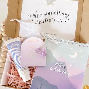 Pode incluir: Uma caixa de presente cheia de papel de seda rosa e uma variedade de itens, incluindo um pequeno frasco de creme para as mãos, um cristal roxo e um caderno com um design de lua e estrelas. O caderno tem o texto "notes & dreams" na capa. A caixa é decorada com uma pequena etiqueta que diz "um pequeno algo só para você".