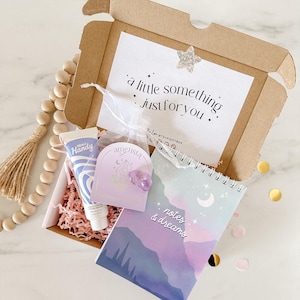 Pode incluir: Uma pequena caixa de cartão cheia de presentes. A caixa contém um caderno roxo e branco com o texto "notas e sonhos", uma pequena bolsa roxa com um cristal dentro, um tubo de creme para as mãos e um bilhete que diz "um pequeno presente só para você".