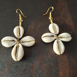Puede incluir: Un par de pendientes hechos a mano con un diseño de cuatro pétalos elaborado con conchas blancas. Las conchas se sujetan con alambre de color dorado, y cada pendiente tiene un gancho de color dorado.