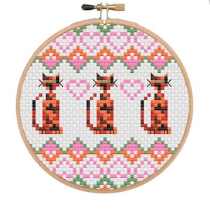 May include: A cross-stitch embroidery hoop featuring three stylized orange and black cats. The cats are positioned below a geometric pattern of pink, green, and orange diamonds. The design is set against a white background.