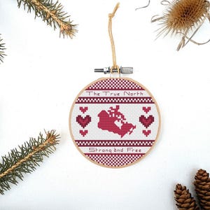 May include: A round wooden ornament with a white background and red stitching. The ornament features a red outline of Canada with red hearts around it. The text "The True North" and "Strong and Free" are stitched in red on the ornament.