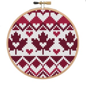 May include: A cross-stitch embroidery hoop featuring a geometric pattern of red and white hearts and maple leaves. The design is set against a white background, with a wooden hoop and a brass adjustment screw.