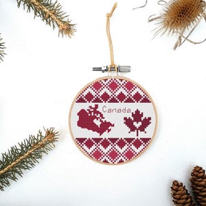 Puede incluir: Un adorno de punto de cruz rojo y blanco con un mapa de Canadá y una hoja de arce. El adorno está enmarcado en un aro de madera y tiene una cuerda para colgar.