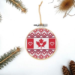 May include: A red and white Canadian flag with a maple leaf and two hearts is embroidered on a round white canvas with a red and white geometric pattern. The canvas is framed in wood and has a string attached for hanging.