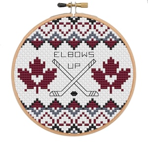 May include: A circular embroidery hoop with a cross-stitch design. The design includes two burgundy maple leaves, crossed hockey sticks, and the text "ELBOWS UP". The background is white with a geometric pattern.