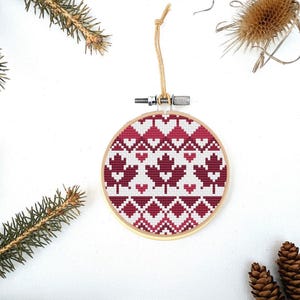 May include: A circular cross-stitch Christmas decoration with a wooden frame. The design showcases a pattern of red maple leaves, hearts, and geometric shapes on a white backdrop. It has a twine loop for hanging.