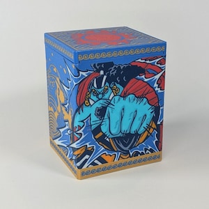 May include: A blue, square box with a detailed illustration of a character with a blue complexion, red hair, and a powerful fist. The box features a red design on the top and a gold wave pattern along the edges. The artwork is in a bold, comic book style.