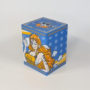 Könnte beinhalten: Eine blaue, würfelförmige Box mit einer handgemalten Illustration eines Charakters mit orangefarbenem Haar und einem gelben Outfit. Die Box hat weiße Tupfen und ein Cartoon-Bild auf der Oberseite. Die Box steht auf einer weißen Oberfläche.