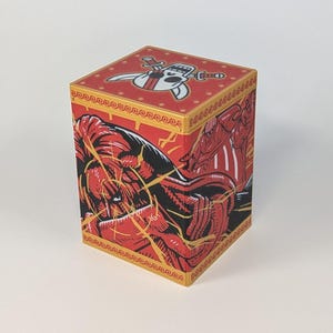 May include: A red and gold cube-shaped box with illustrations. The box features a cartoon character with a sword on the top and a red and black illustration of a figure on the sides. The box has a gold border.