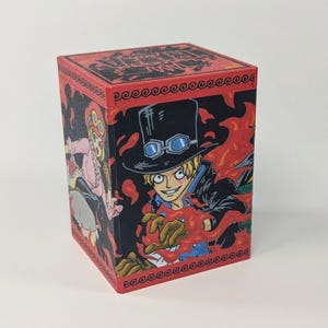 May include: A red and black box featuring anime characters. The box has a red border with a repeating wave pattern. The sides of the box feature illustrations of characters with flames and other design elements.