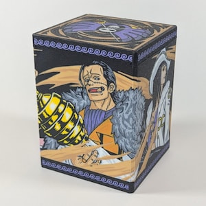 Può includere: Una scatola nera quadrata per riporre carte con illustrazioni di personaggi della serie anime One Piece. La scatola ha un design dettagliato con un personaggio su ogni lato e un bordo decorativo. La parte superiore della scatola ha un design viola e oro.