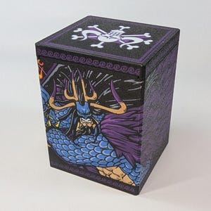 Puede incluir: Una caja de almacenamiento rectangular con base negra y una ilustración gráfica en morado y azul de un personaje con cuernos en el frente. La tapa muestra un emblema blanco de calavera y tibias cruzadas. La caja tiene un acabado texturizado.