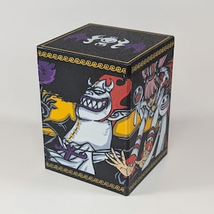 May include: Black, cube-shaped storage box featuring colorful anime-style illustrations. The box has a gold border and a purple design on the top. The artwork includes a white character with a large grin, red hair, and a yellow background.