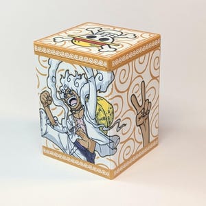May include: A square storage box featuring a tan and white spiral pattern. The top lid displays a skull and crossbones logo with a straw hat. The front panel shows a cartoon character with white hair, a white shirt, and a yellow straw hat.