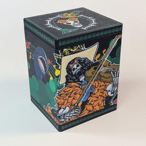 May include: Black cube-shaped box featuring a cartoon skeleton playing a violin, with a shark and musical notes. The top of the box has a skull wearing sunglasses and a crown. The box is decorated with green, orange, and yellow accents.