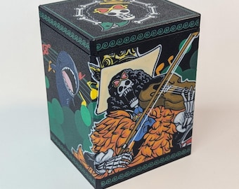 Puede incluir: Caja negra cúbica con un esqueleto de dibujos animados tocando el violín, un tiburón y notas musicales. La parte superior de la caja tiene una calavera con gafas de sol y una corona. La caja está decorada con detalles verdes, naranjas y amarillos.