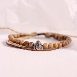 May include: A multi-strand bracelet featuring a silver Buddha charm and natural stone beads in shades of brown and tan. The bracelet is made with a woven cord and has a neutral, earthy aesthetic. The beads are approximately 8mm in diameter.