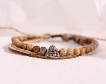 May include: A multi-strand bracelet featuring a silver Buddha charm and natural stone beads in shades of brown and tan. The bracelet is made with a woven cord and has a neutral, earthy aesthetic. The beads are approximately 8mm in diameter.