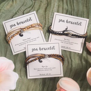 Pode incluir: São apresentados três conjuntos de "Zen Bracelet". Cada pulseira é apresentada com um cartão que detalha o tipo de pedra: Picture Jasper, Lava Stone e Olho de Tigre. As pulseiras têm contas de madeira e um pequeno pingente, com os cartões que dizem "Handmade with love."
