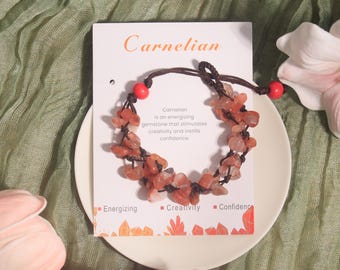 May include: A carnelian gemstone bracelet with a brown cord and red beads. The bracelet is displayed on a white plate with a card that reads "Carnelian" and describes the gemstone's properties. The card also has the words "Energizing", "Creativity", and "Confidence".