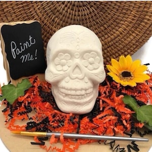 May include: A white ceramic skull with embossed floral and decorative patterns, accompanied by a small black chalkboard sign with the white text Paint me!, a yellow sunflower, green leaves, and a silver paintbrush on orange and black paper shreds.