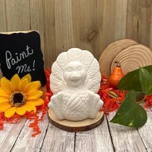 May include: White ceramic turkey figurine with a happy face, ready to be painted. The figurine is sitting on a wooden base and surrounded by orange and brown fall-themed decorations. A small chalkboard sign reads "Paint Me!"