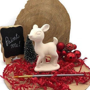Puede incluir: Figurita de ciervo de cerámica blanca con un letrero de "píntame" y un pincel. El ciervo está rodeado de decoraciones navideñas rojas y doradas.